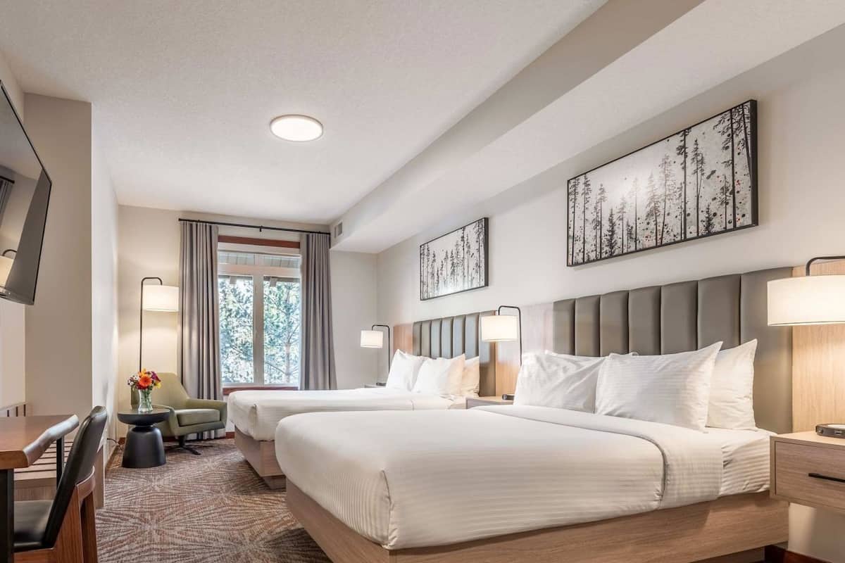 A hotel room with a large bed, a desk, a chair, a small table with a vase, and a window with curtains.