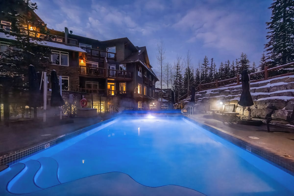 A heated outdoor pool with a stone border, surrounded by a multi-story building with balconies and a snow-covered slope.