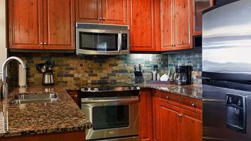 A kitchen with wooden cabinets, a stainless steel microwave, a stove, a refrigerator, granite countertops, and a stone backsplash.