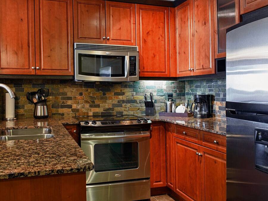 A kitchen with wooden cabinets, a stainless steel microwave, a stove, a refrigerator, granite countertops, and a stone backsplash.