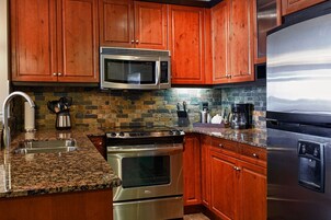 A kitchen with wooden cabinets, a stainless steel microwave, a stove, a refrigerator, granite countertops, and a stone backsplash.