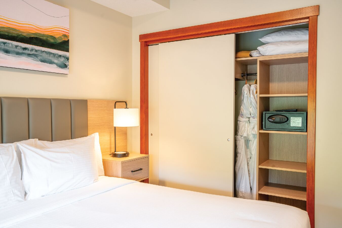 Habitación de hotel con cama, mesita de noche con lámpara, armario de madera y un cuadro en la pared.