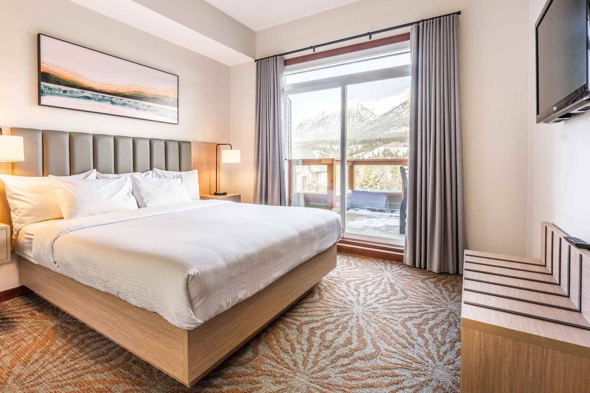 A hotel room with a large bed, a flat-screen TV, a painting above the bed, and a view of a snowy landscape through the window.