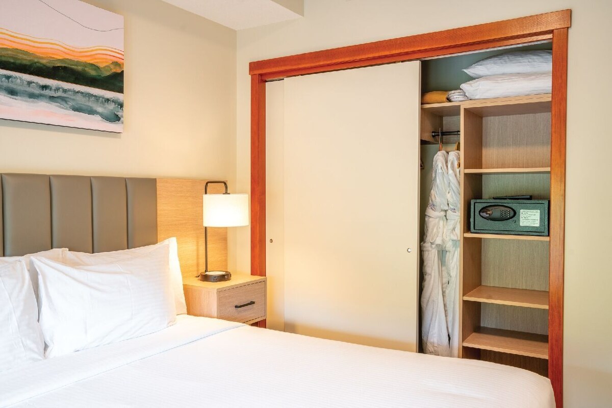 Habitación de hotel con cama, mesita de noche con lámpara, armario de madera y un cuadro en la pared.