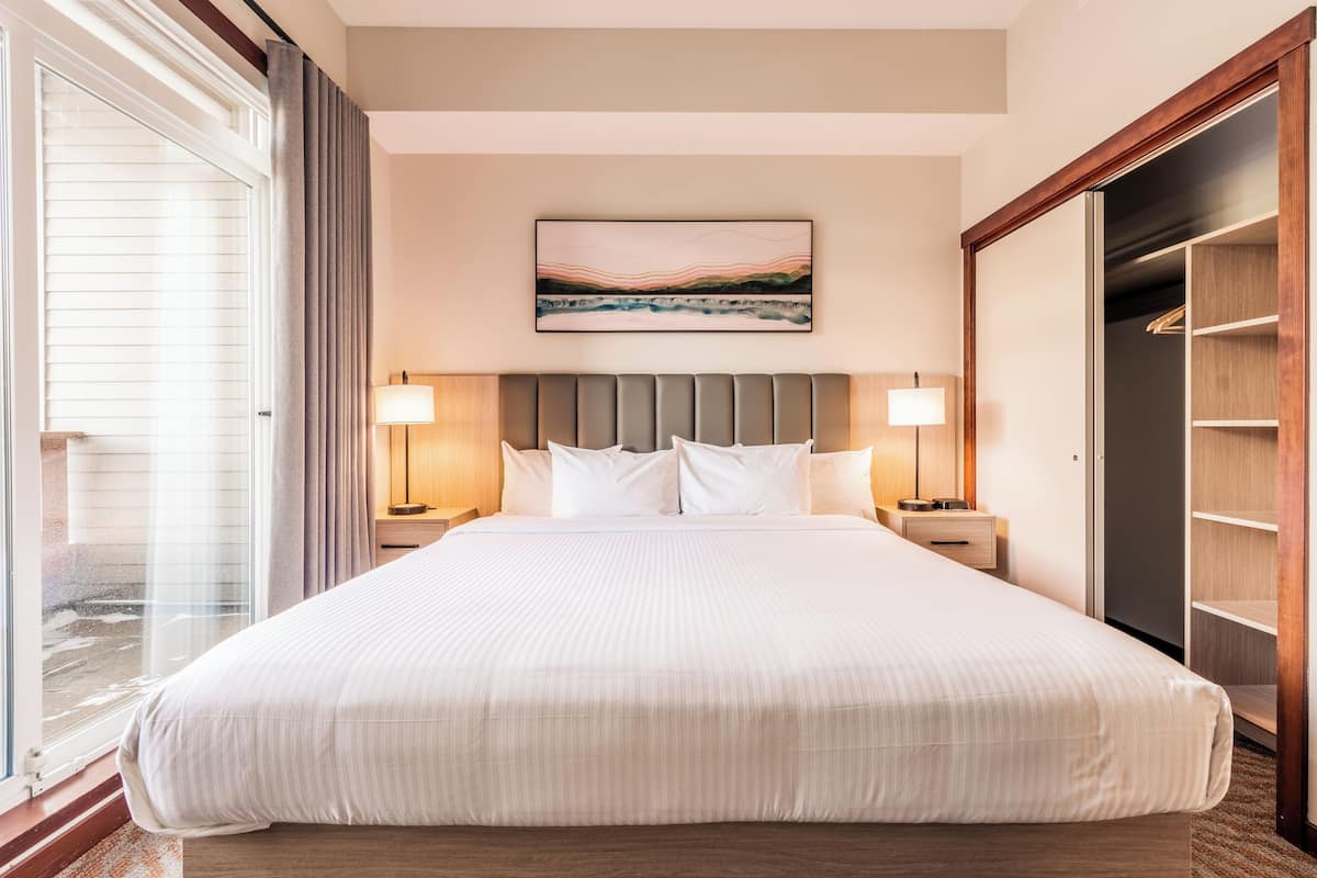 A large bed with a headboard, two bedside tables with lamps, a large window with curtains, and a closet with shelves.