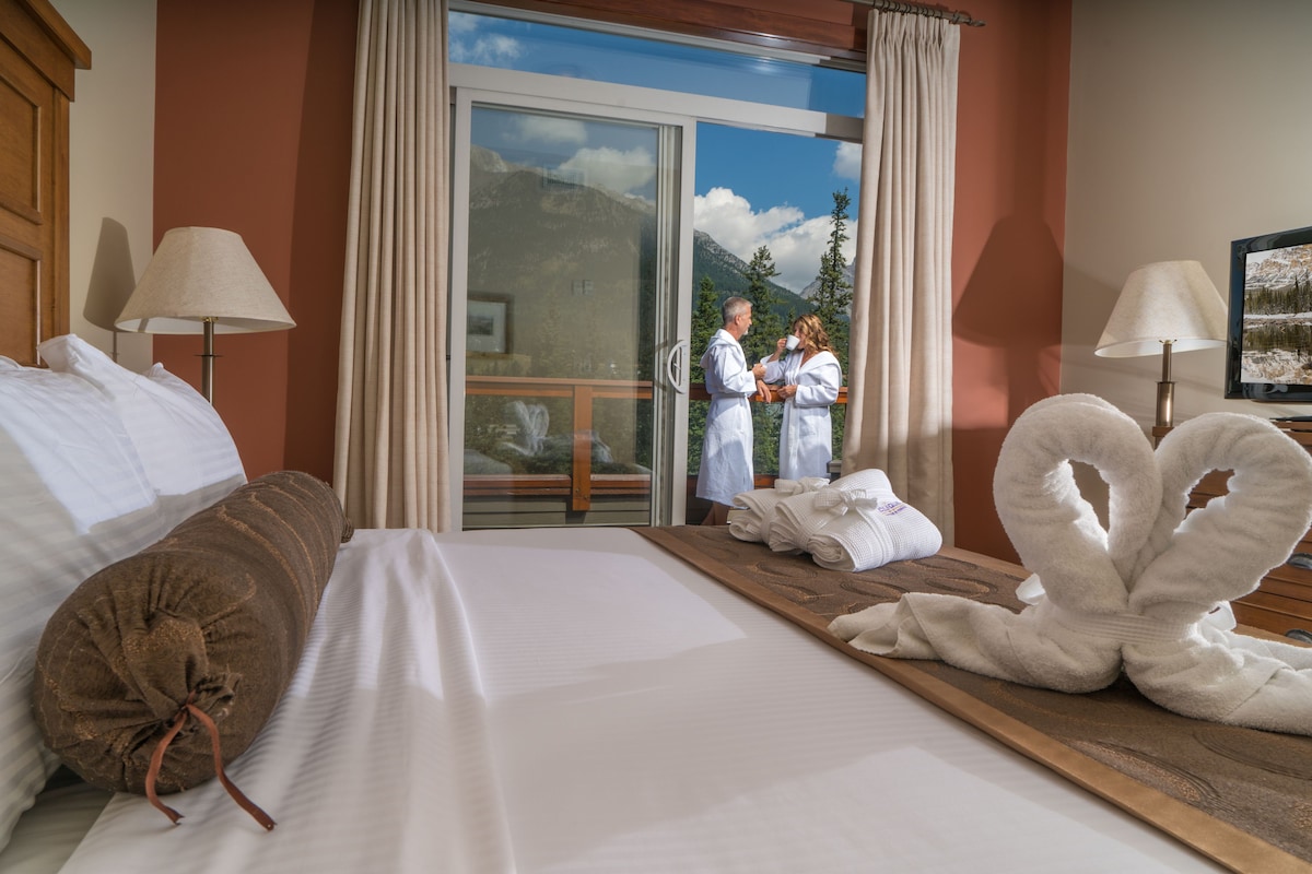 Habitación de hotel con una cama, cabecero de madera, un ventanal y vistas a las montañas.