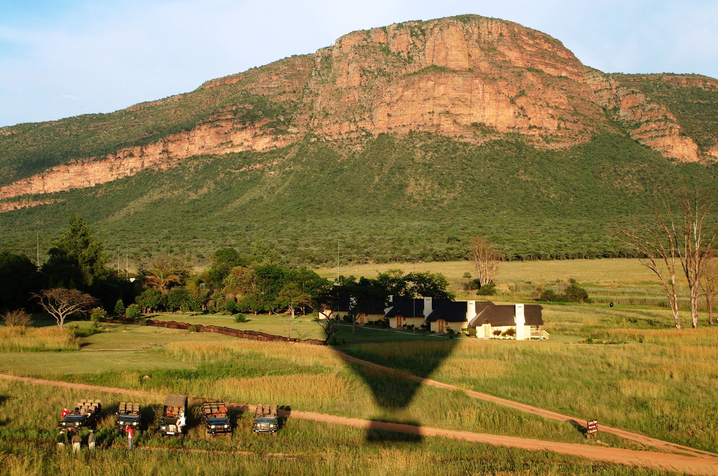 A rural landscape with a mountain, a dirt road, and several vehicles parked near a building with a white facade.
