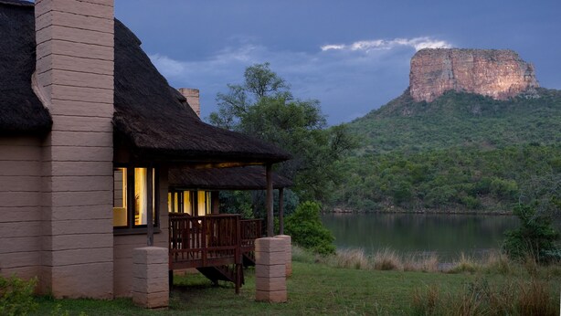 A lodge with a thatched roof, a stone chimney, and a view of a lake and a large rock formation. - Entabeni Mountain Lodges (Mookgopong)