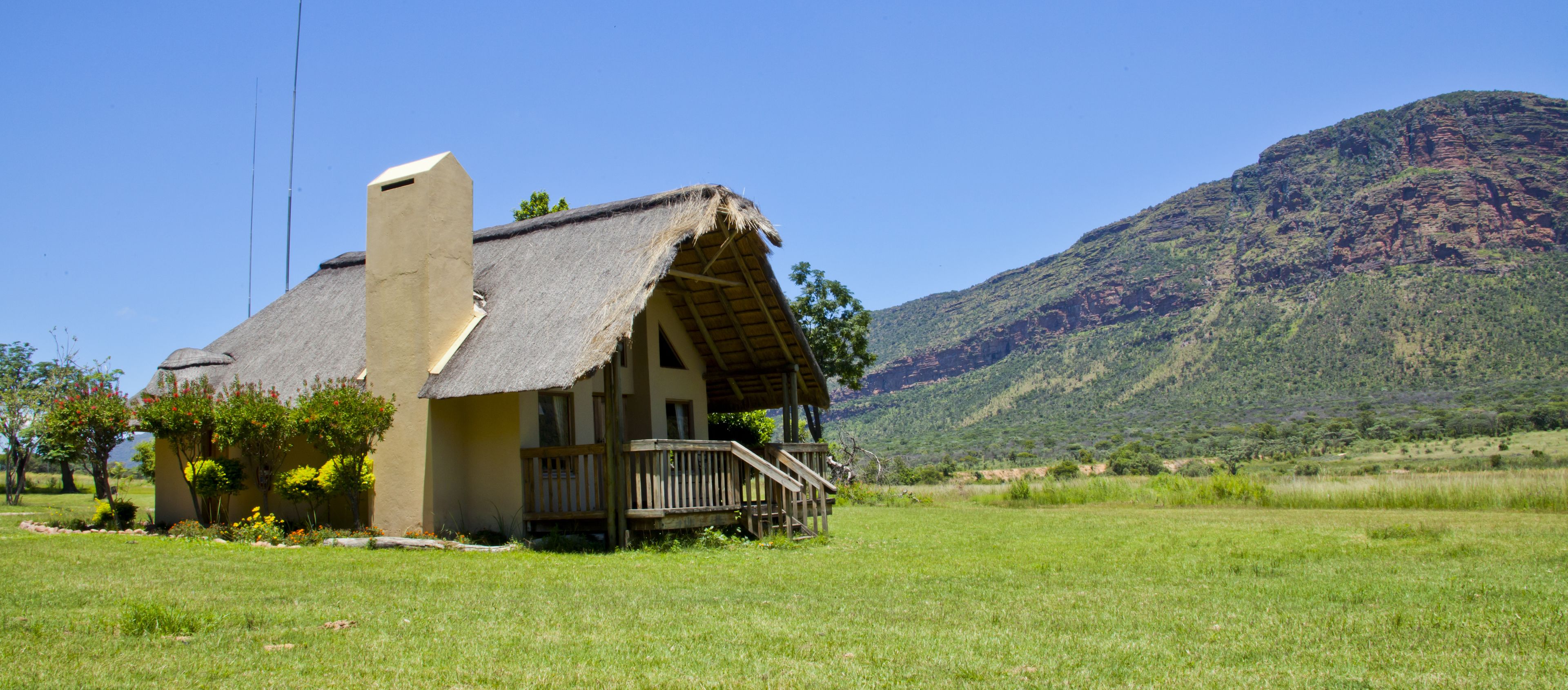 A rustic house with a thatched roof, a wooden porch, and a chimney. It is situated in a grassy field with a mountain in the background.