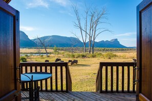 An open wooden door leading to a deck with a table and chairs, overlooking a grassy field with animals and mountains in the background.