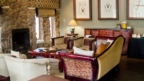 A rustic interior with a stone fireplace, wooden ceiling beams, and a chandelier. There are framed pictures on the wall and a wicker bench. - Entabeni Mountain Lodges (Mookgopong)