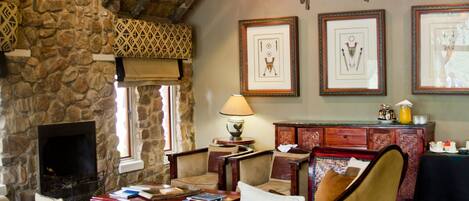 A rustic interior with a stone fireplace, wooden ceiling beams, and a chandelier. There are framed pictures on the wall and a wicker bench.