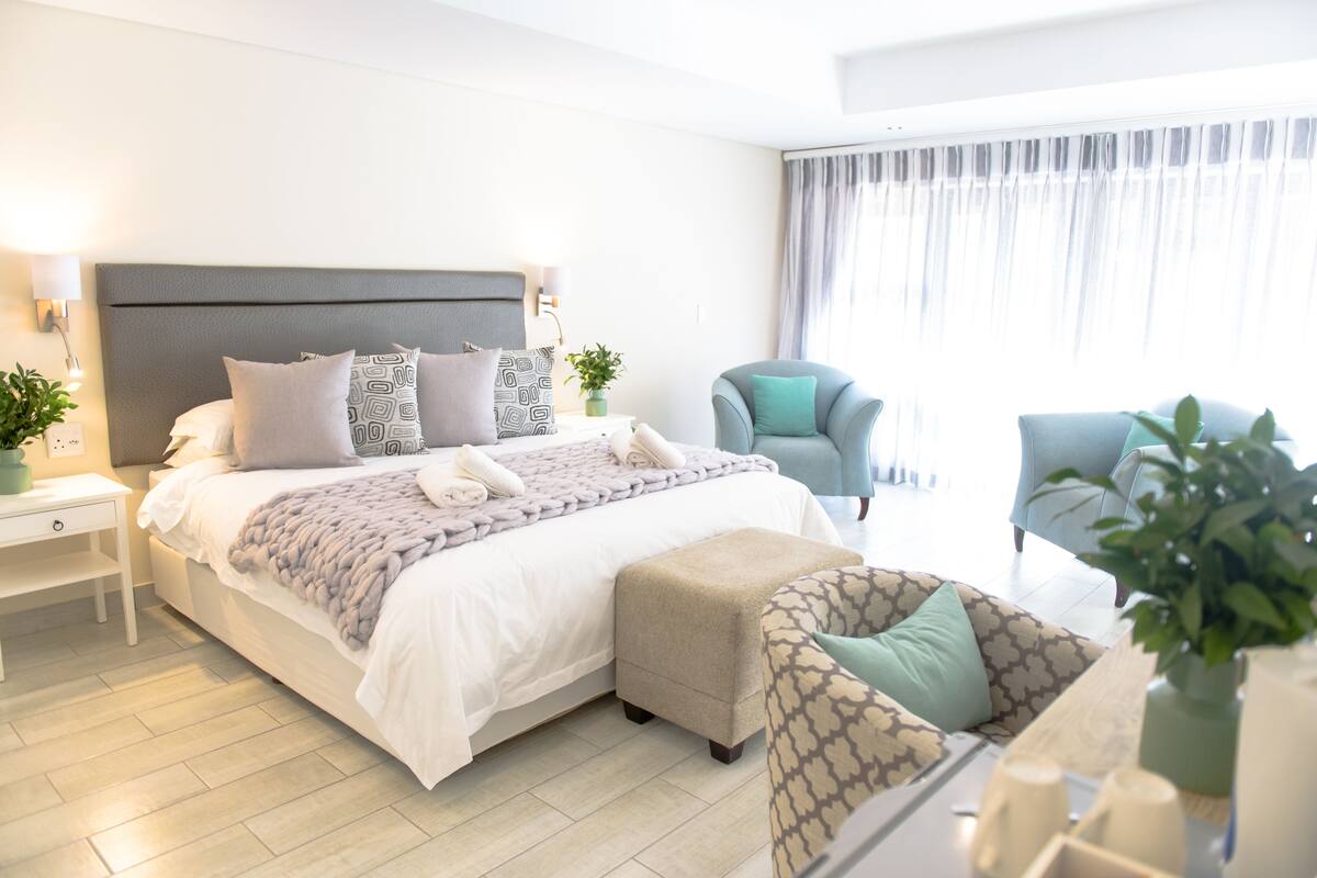 A modern hotel room with a large bed, a sitting area with a chair, a small table, and a potted plant.