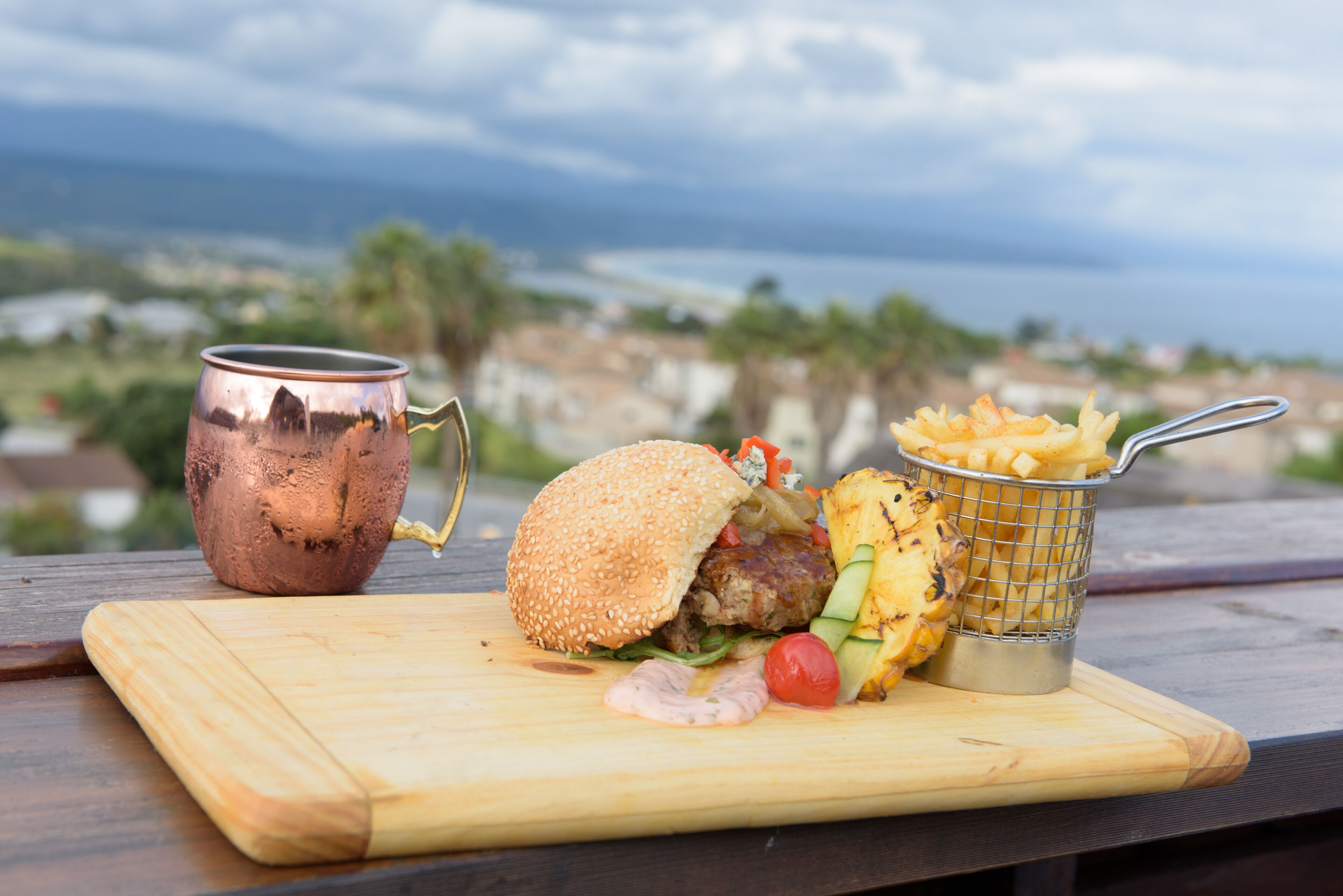 A meal of a hamburger, fries, and a drink on a wooden board with a scenic view in the background.