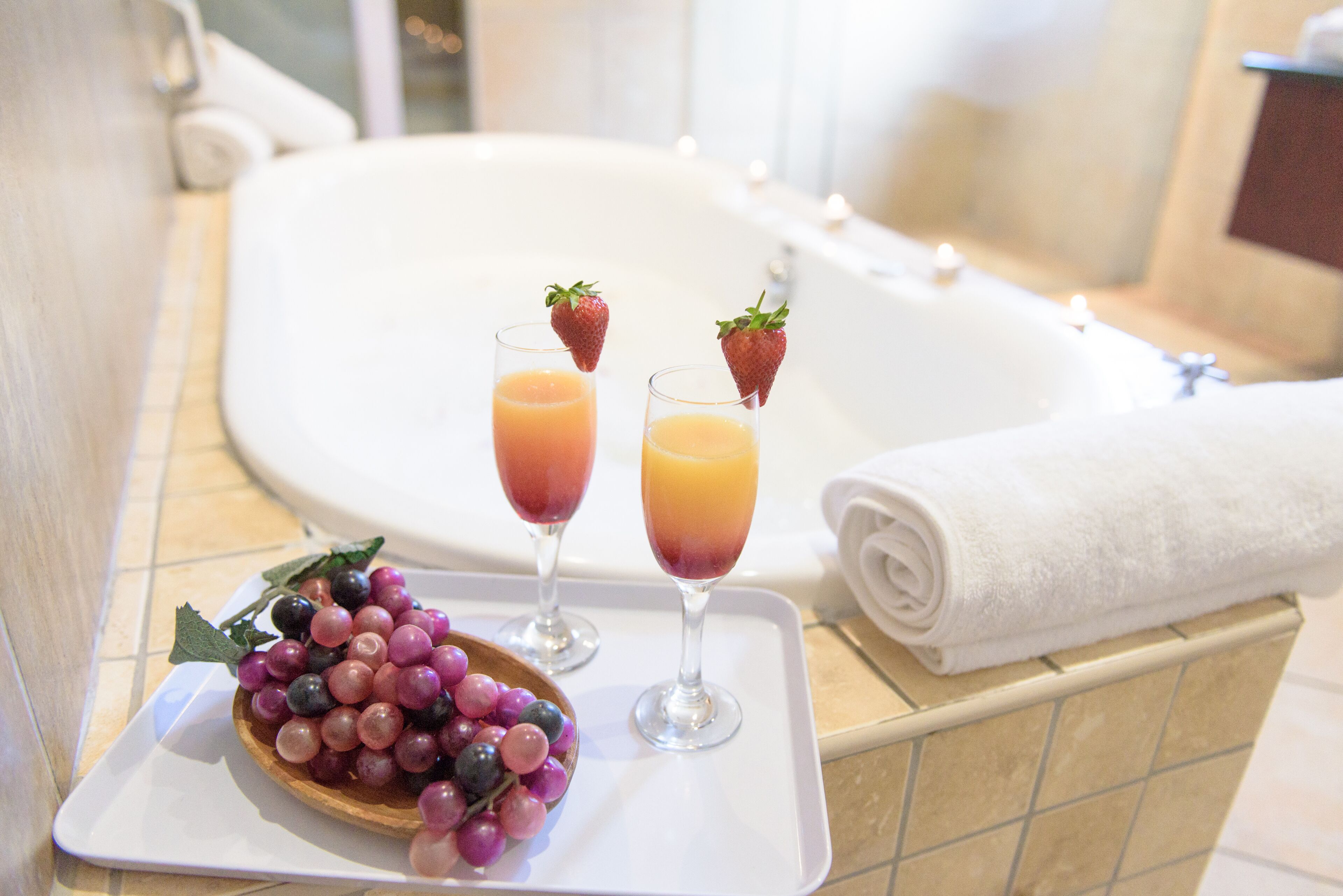 A bathroom with a bathtub, two glasses of fruit juice, a bowl of berries, and a rolled towel.
