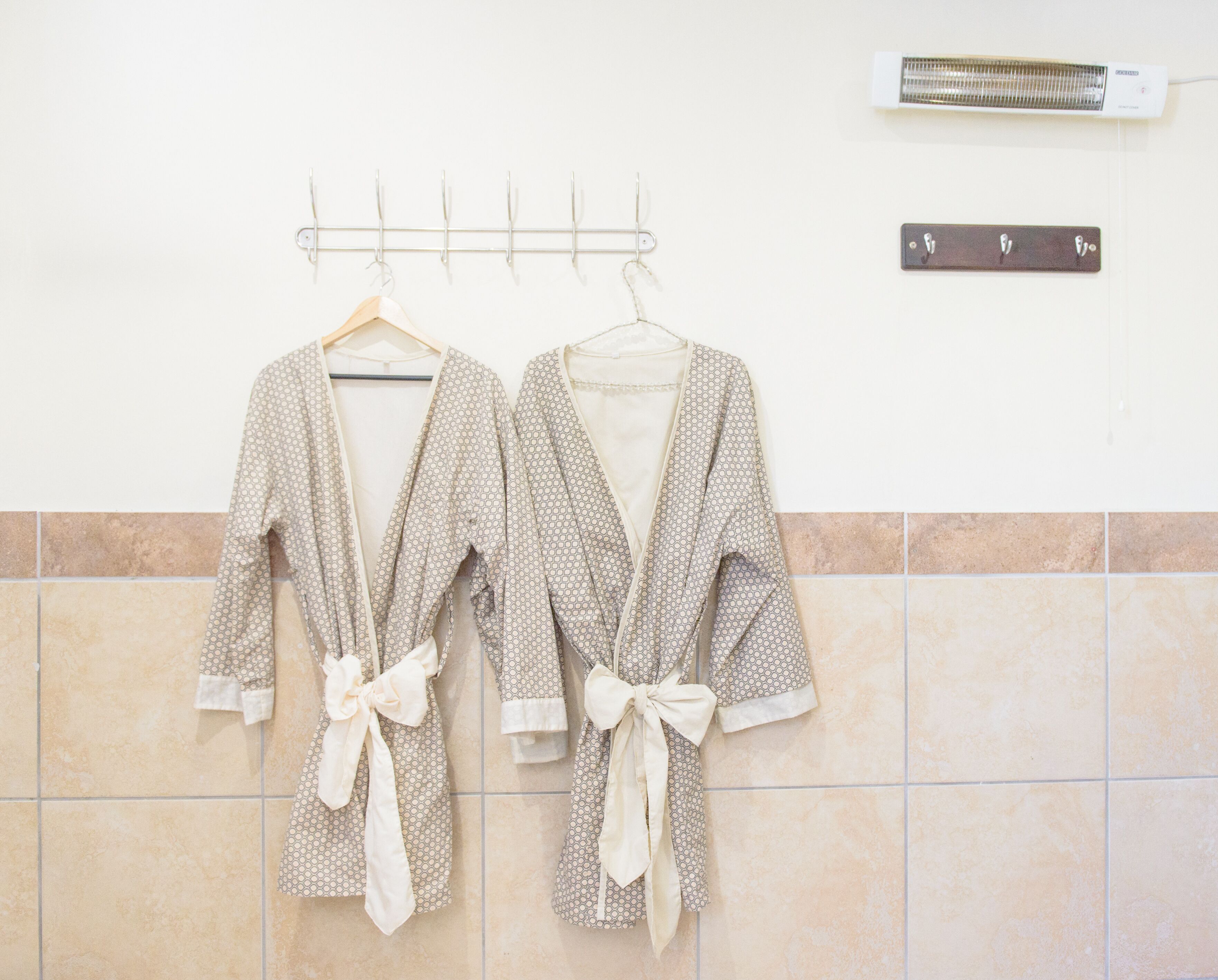 Zwei gemusterte Bademäntel mit Schleifen hängen an einem Gestell vor einer gefliesten Wand, darüber befinden sich ein Heizkörper und eine Fußmassagegerät.