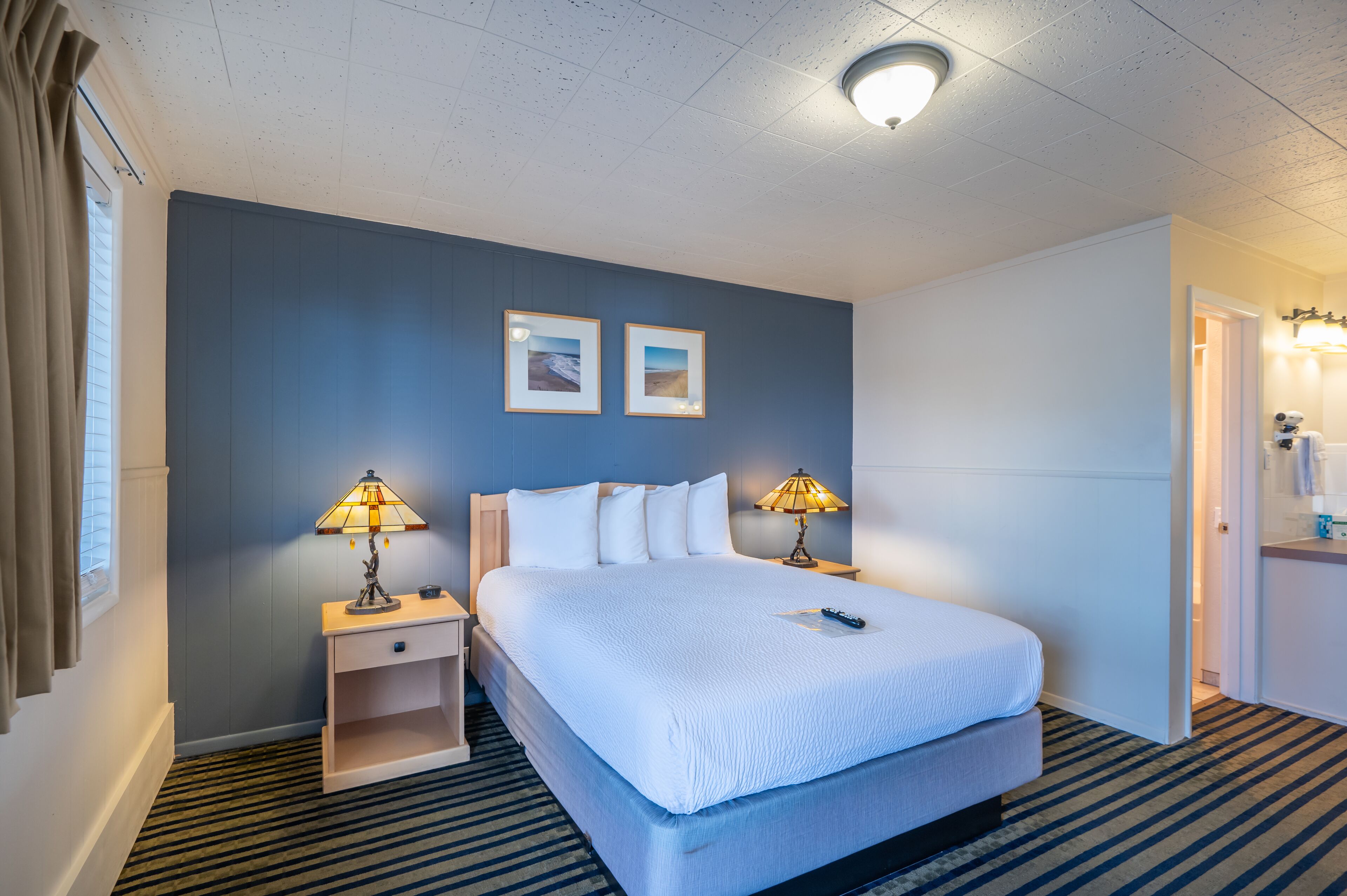 A hotel room with a large bed, two bedside lamps, a nightstand, a dresser, and a door leading to a bathroom.