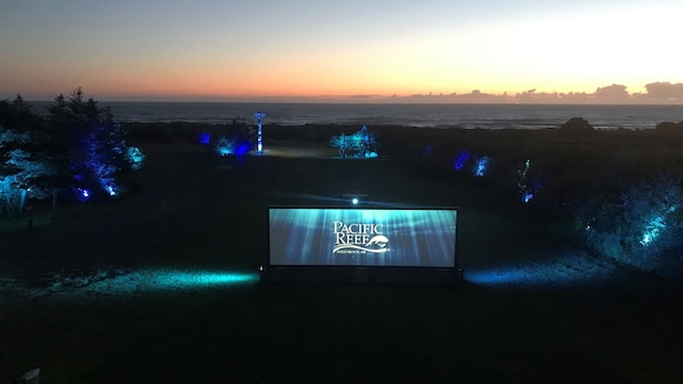 A large outdoor screen displaying "Pacific Reef" with a view of the ocean and trees illuminated by blue lights. - Pacific Reef Hotel & Light Show (Gold Beach)