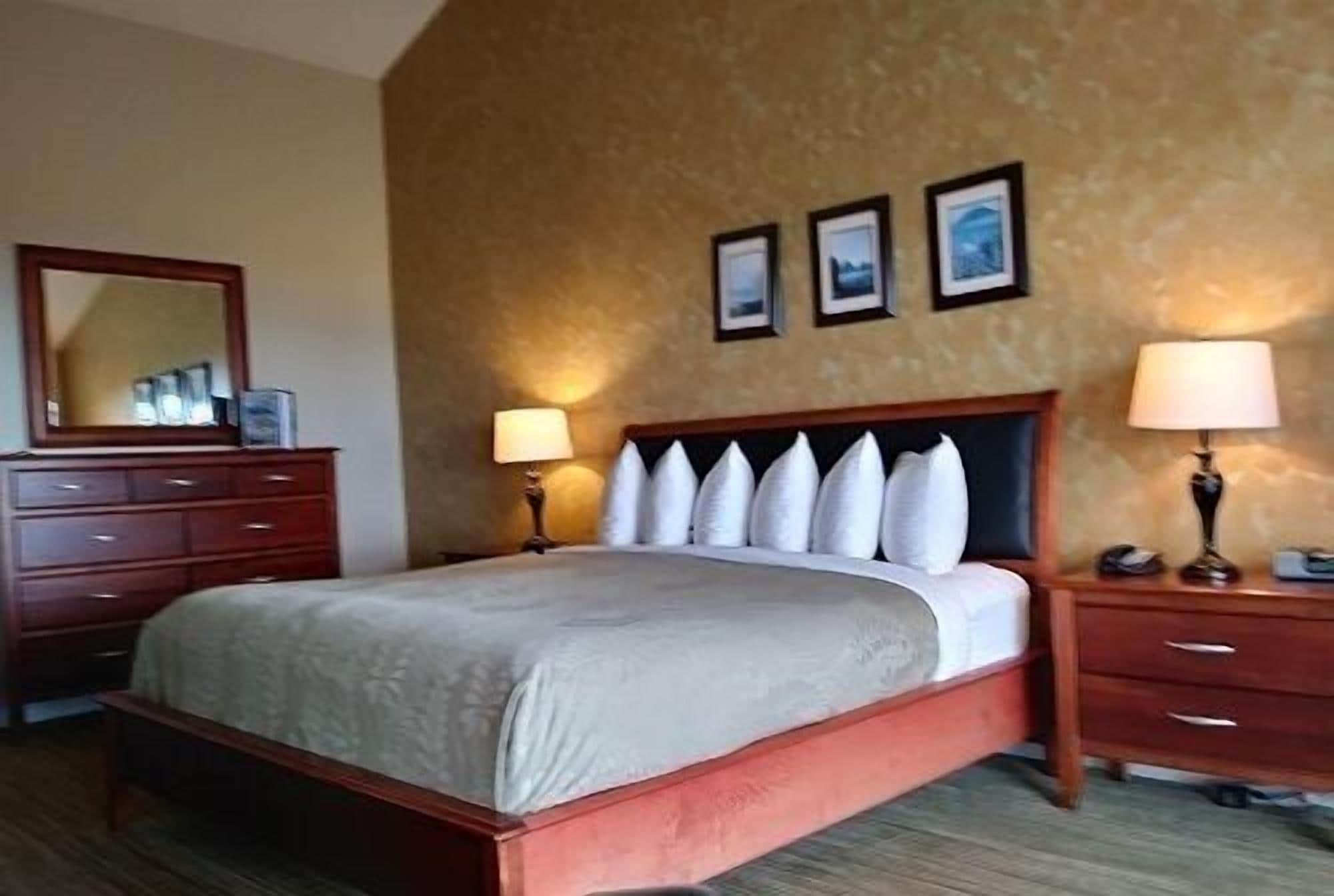 Una habitación de hotel con una cama grande, cabecera de madera, dos mesitas de noche con lámparas, un aparador y tres cuadros enmarcados en la pared.