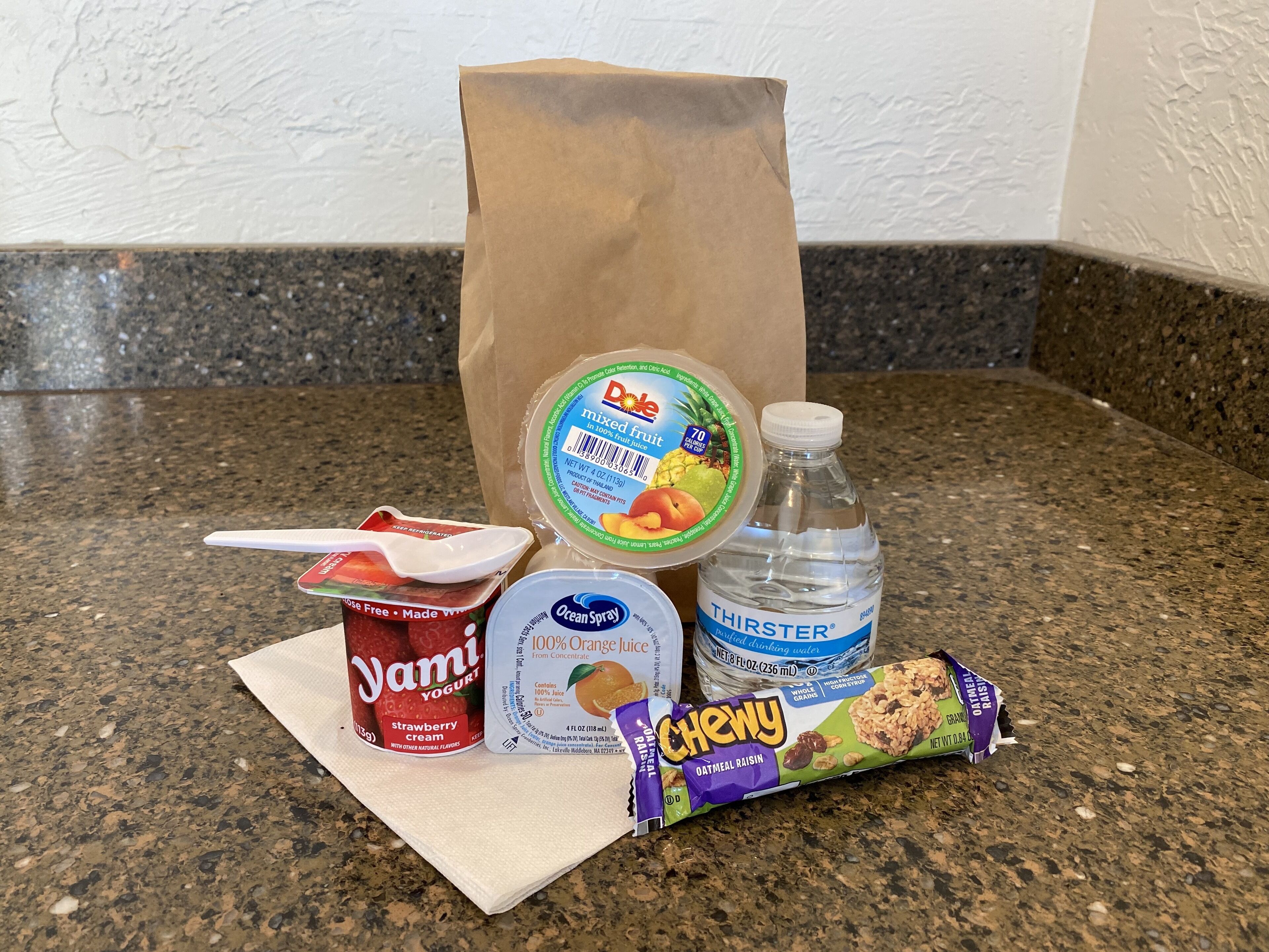 A variety of food items including yogurt, a bottle of water, a bag of chips, and a snack bar on a kitchen counter.