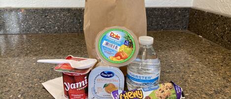 A variety of food items including yogurt, a bottle of water, a bag of chips, and a snack bar on a kitchen counter.