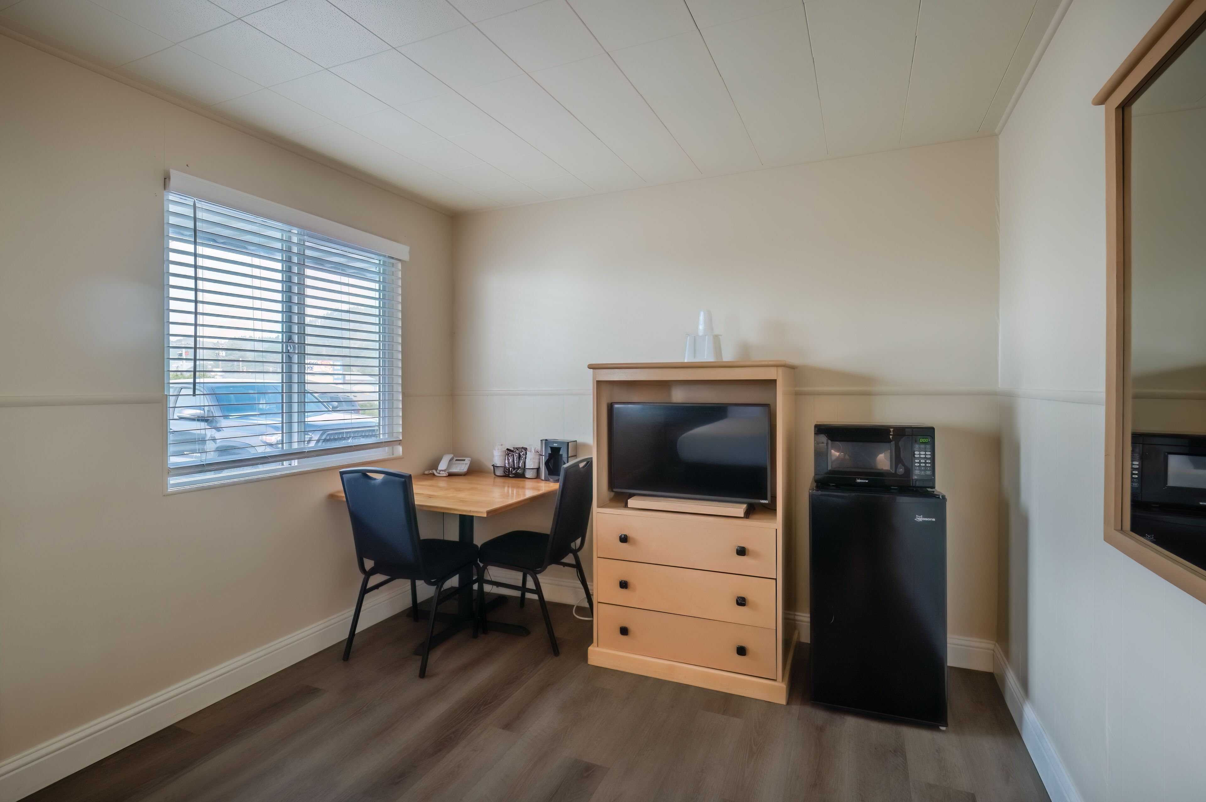 Een compacte kamer met een houten bureau, twee zwarte stoelen, een zwarte koelkast, een magnetron en een raam met jaloezieën.