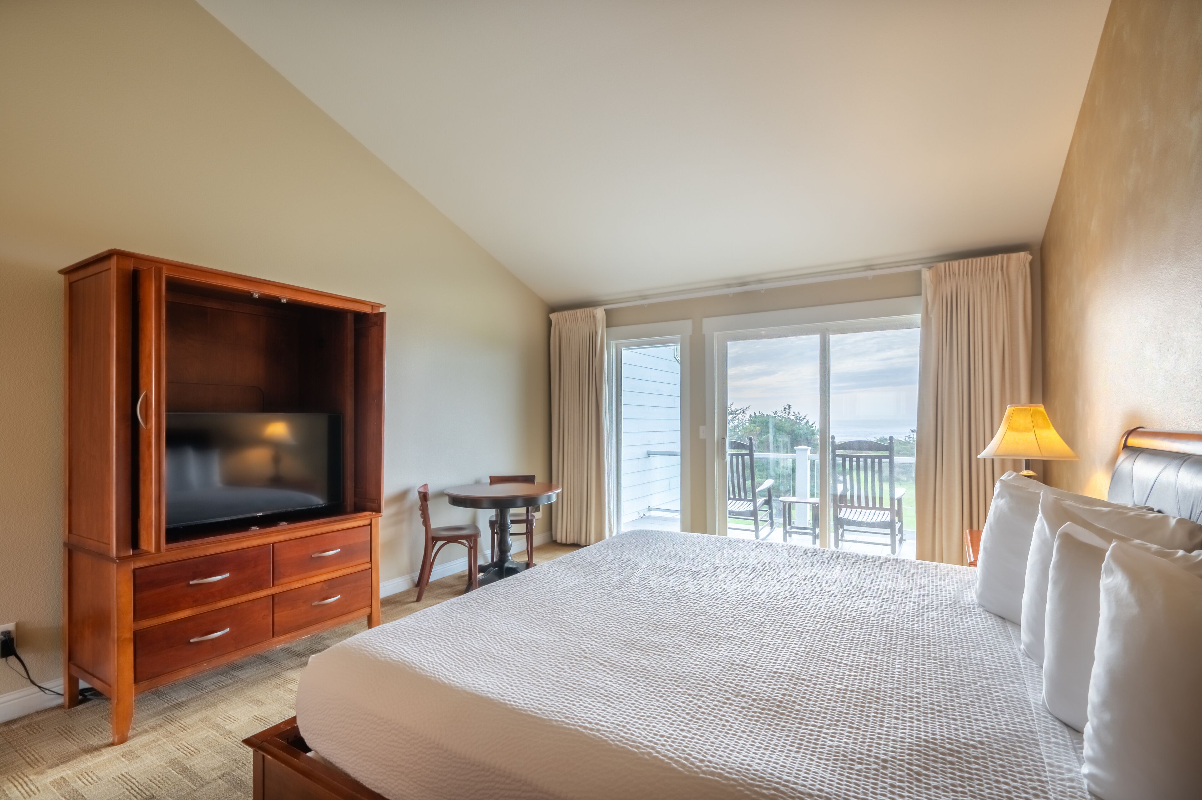 A hotel room with a large bed, wooden TV stand, a small table with a chair, and a balcony with curtains.