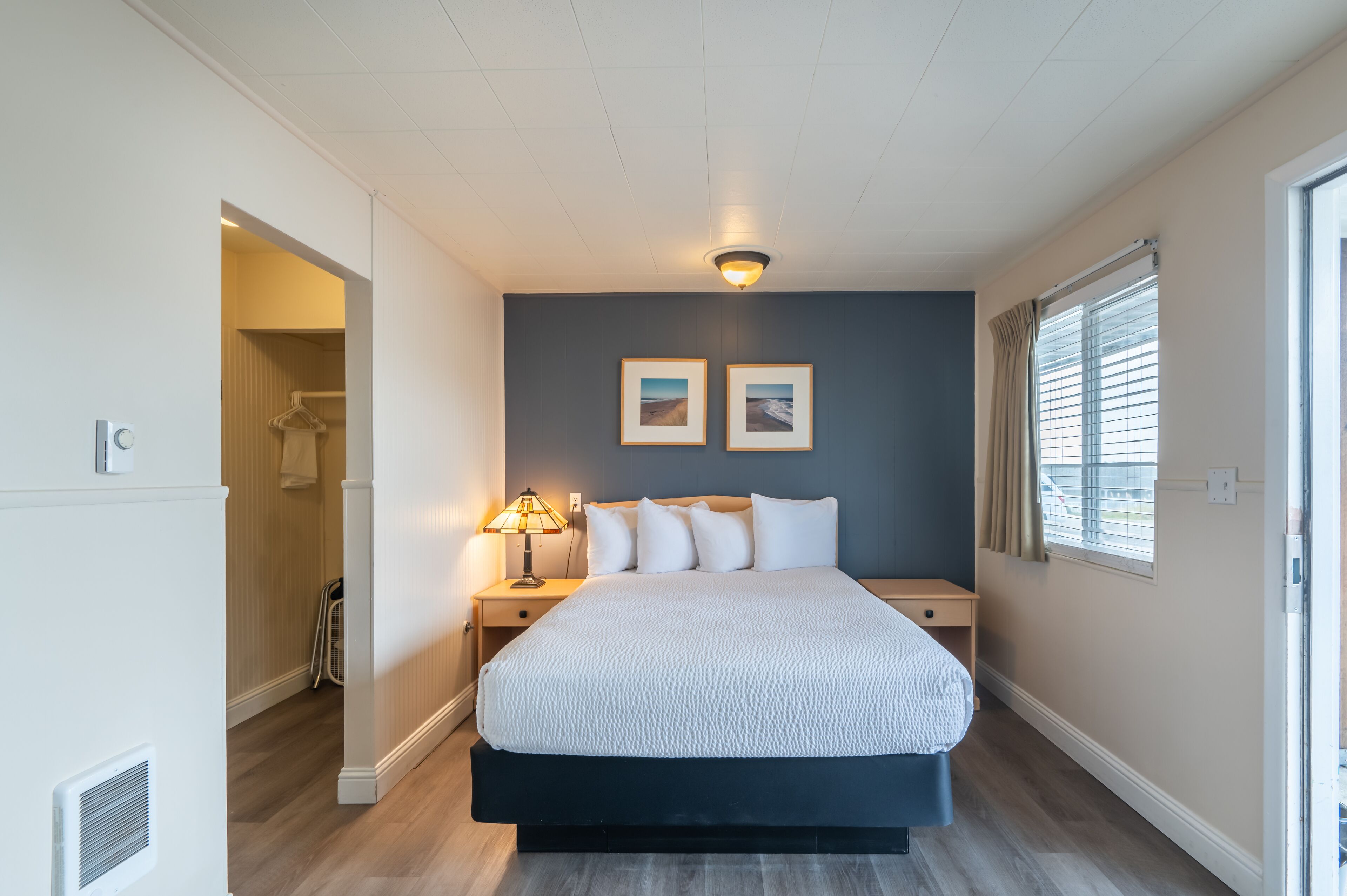A bedroom with a bed, two bedside tables, a nightstand, a wall-mounted light fixture, and a window with blinds.