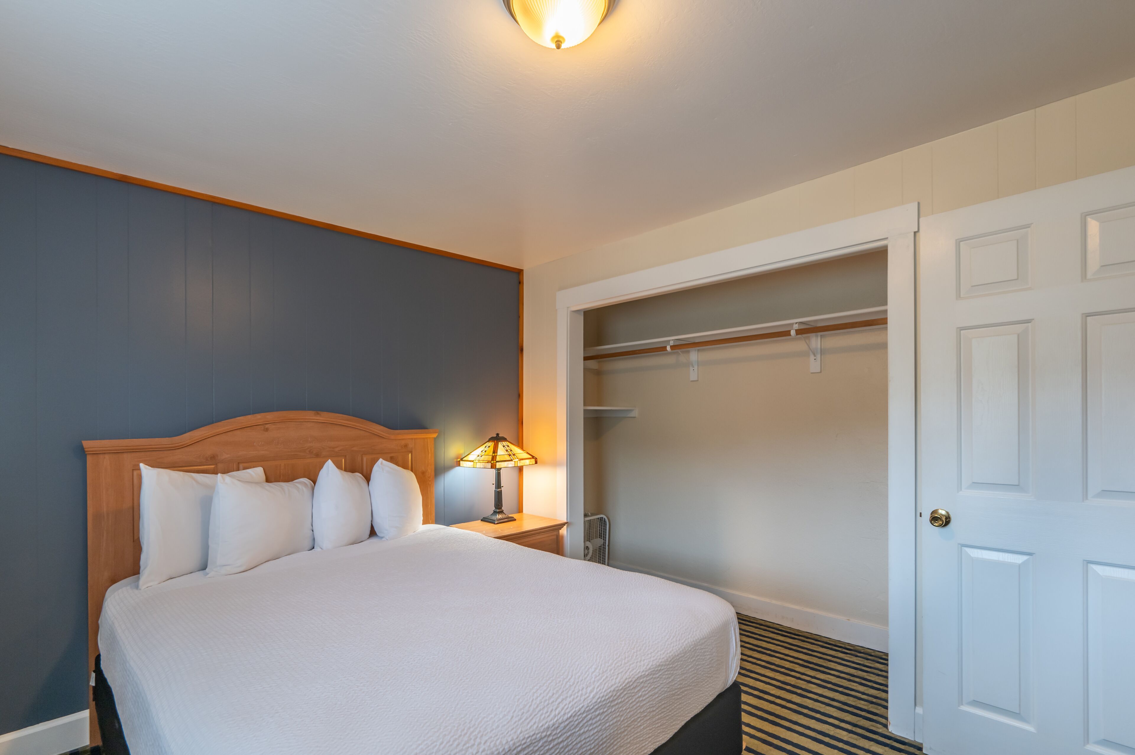 Quarto de hotel com cama grande, mesa de cabeceira com abajur, closet espaçoso e uma porta.