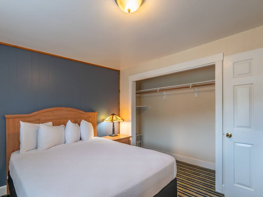 Quarto de hotel com cama grande, mesa de cabeceira com abajur, closet espaçoso e uma porta.