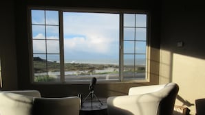 Ein Zimmer mit großem Fenster und Blick auf eine Küstenlandschaft, zwei weiße Stühle und ein kleiner Tisch mit einem Mikrofon.