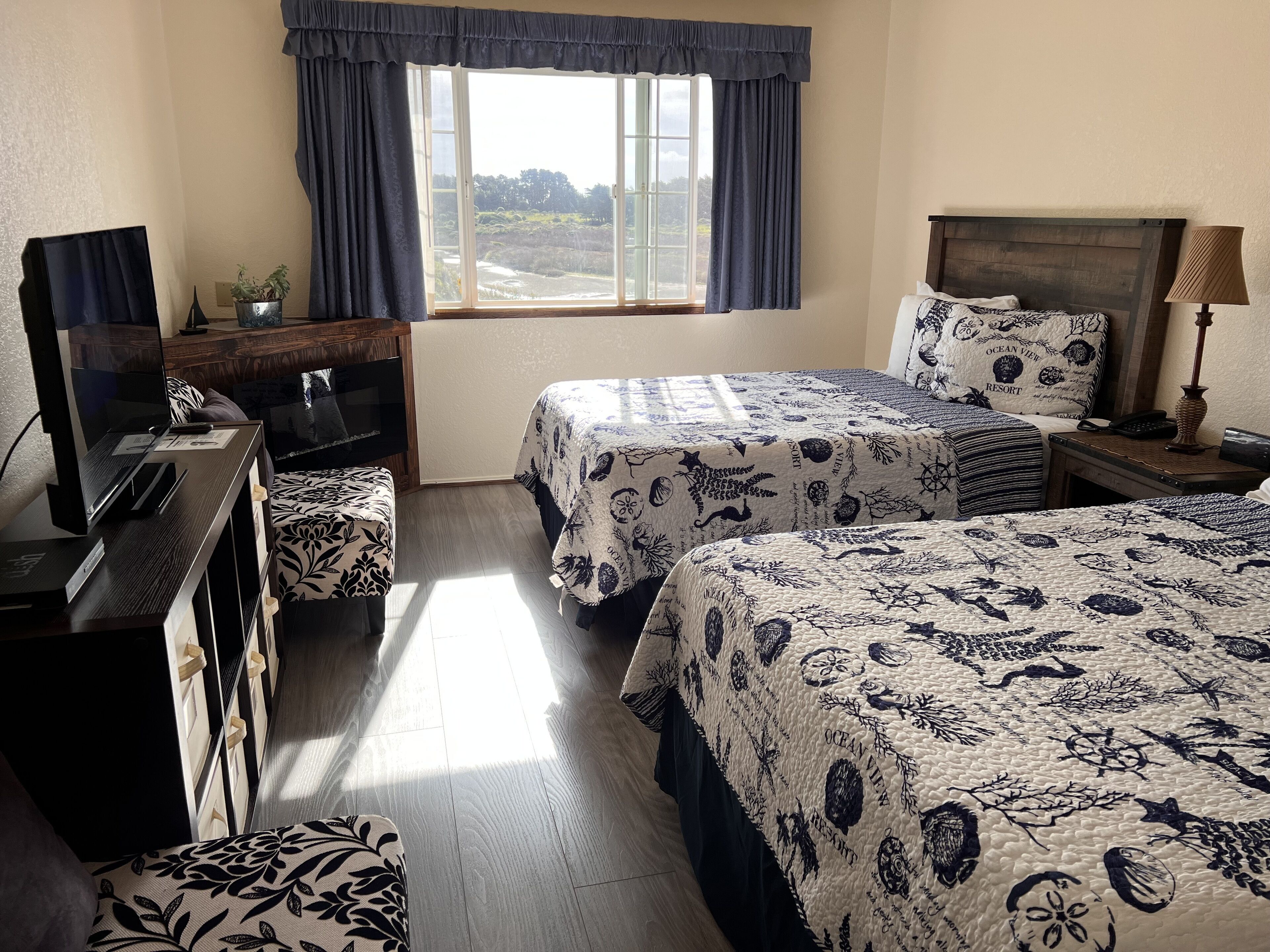 Habitación de hotel con dos camas, un escritorio, un televisor y un ventanal con cortinas.