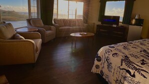 Ein Hotelzimmer mit großem Fenster und Blick auf Meer und Hügellandschaft, ausgestattet mit einer Couch, einem Sessel, einem kleinen Tisch, einem Bett mit gemustertem Überwurf und einem Fernseher auf einem Schrank.