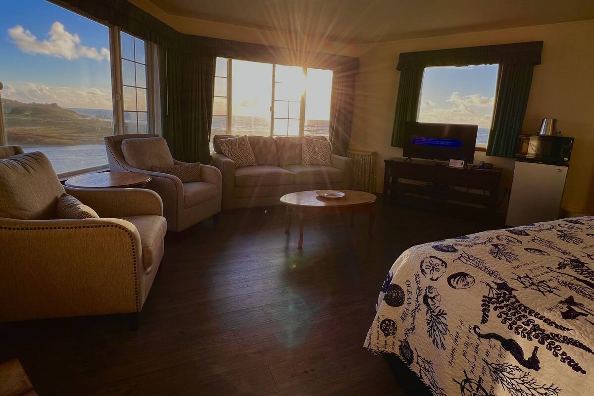 Ein Hotelzimmer mit großem Fenster und Blick auf Meer und Hügellandschaft, ausgestattet mit einer Couch, einem Sessel, einem kleinen Tisch, einem Bett mit gemustertem Überwurf und einem Fernseher auf einem Schrank.