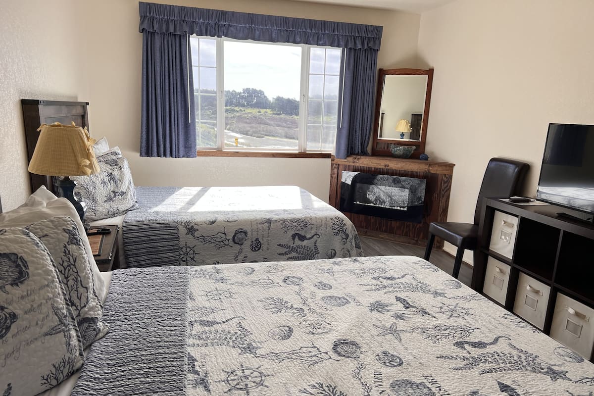 Ein Hotelzimmer mit zwei Betten, einem Schreibtisch mit Fernseher, einem Fenster mit Vorhängen und einem Spiegel.