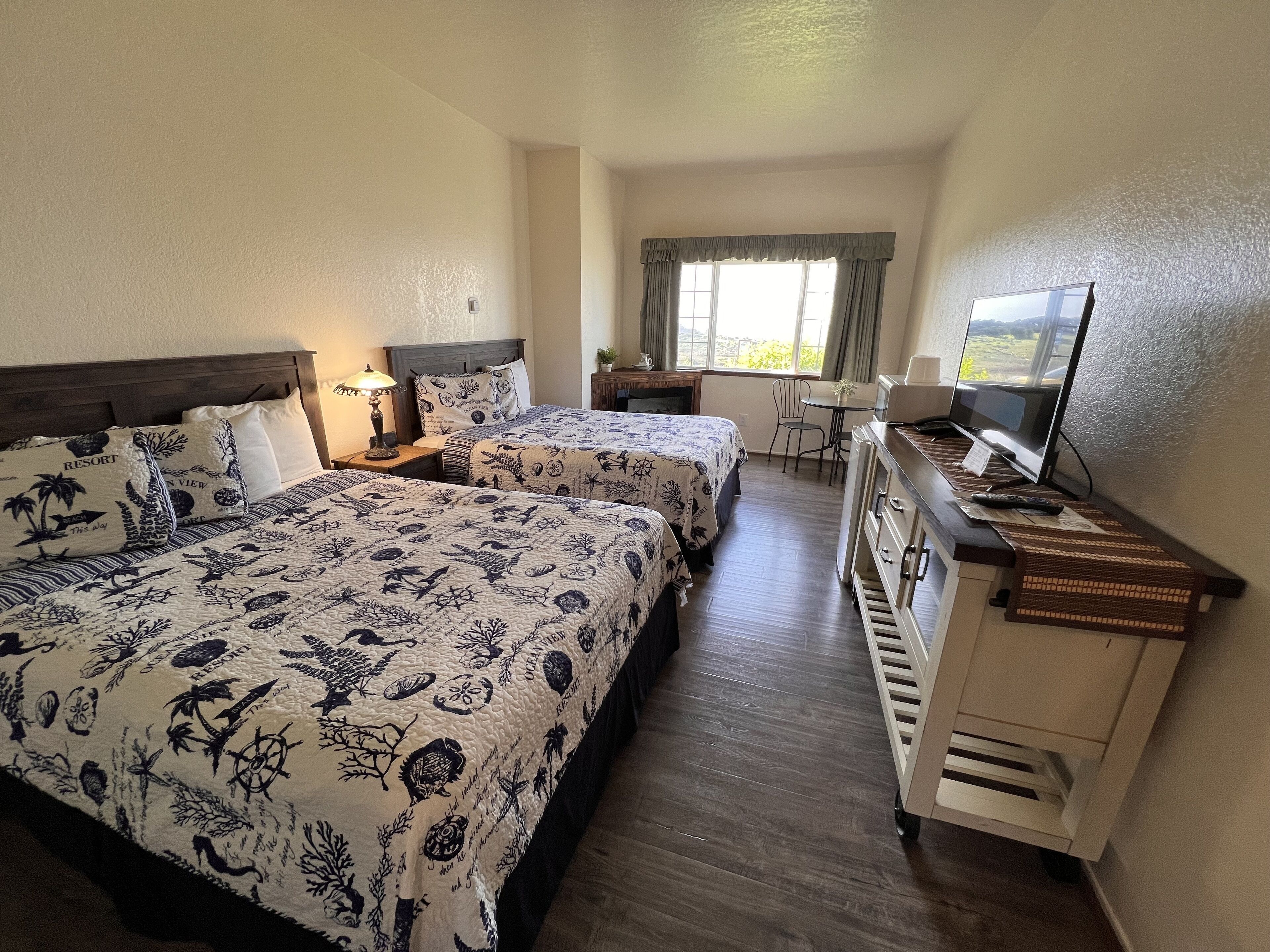 A hotel room with two beds, a desk, a TV, and a window with curtains.