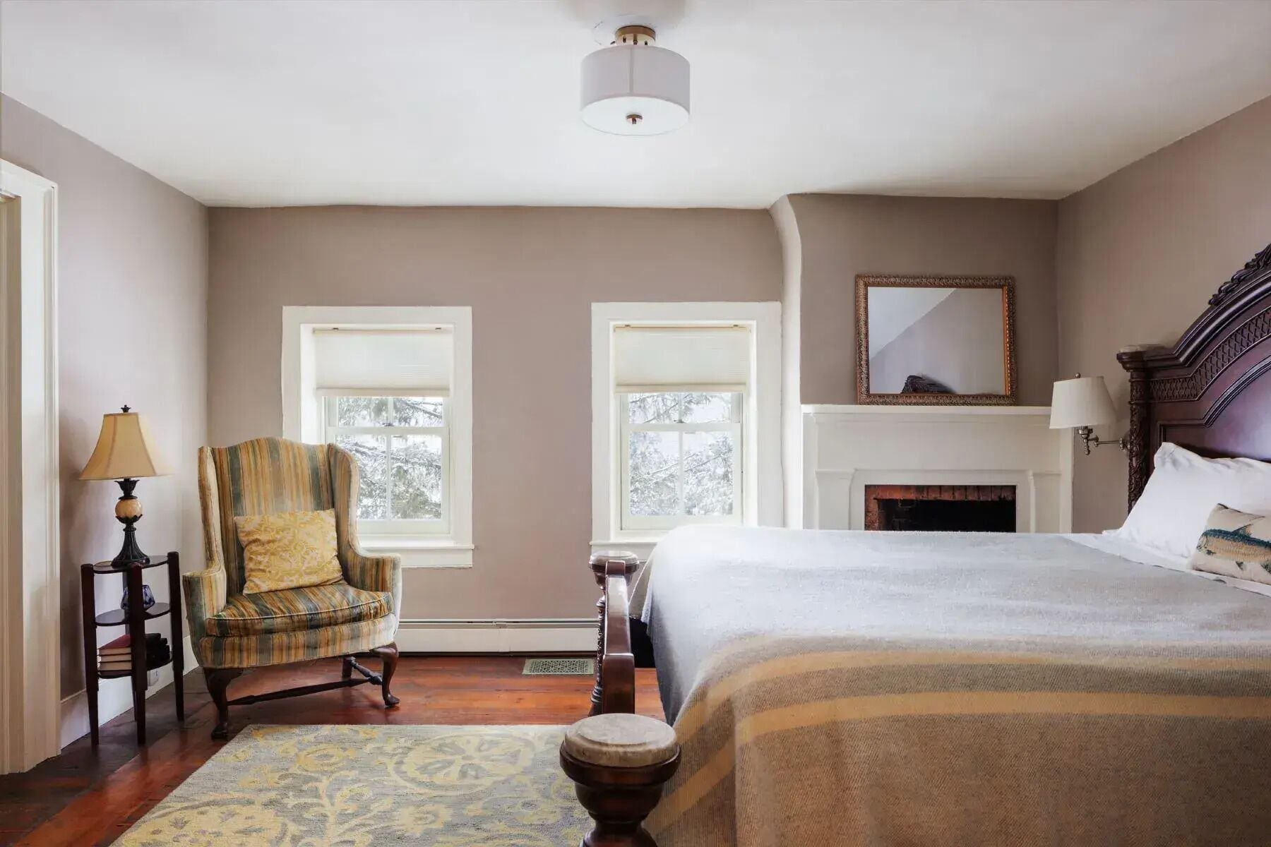A bedroom with a large bed, two windows, a fireplace, a chair, and a lamp.