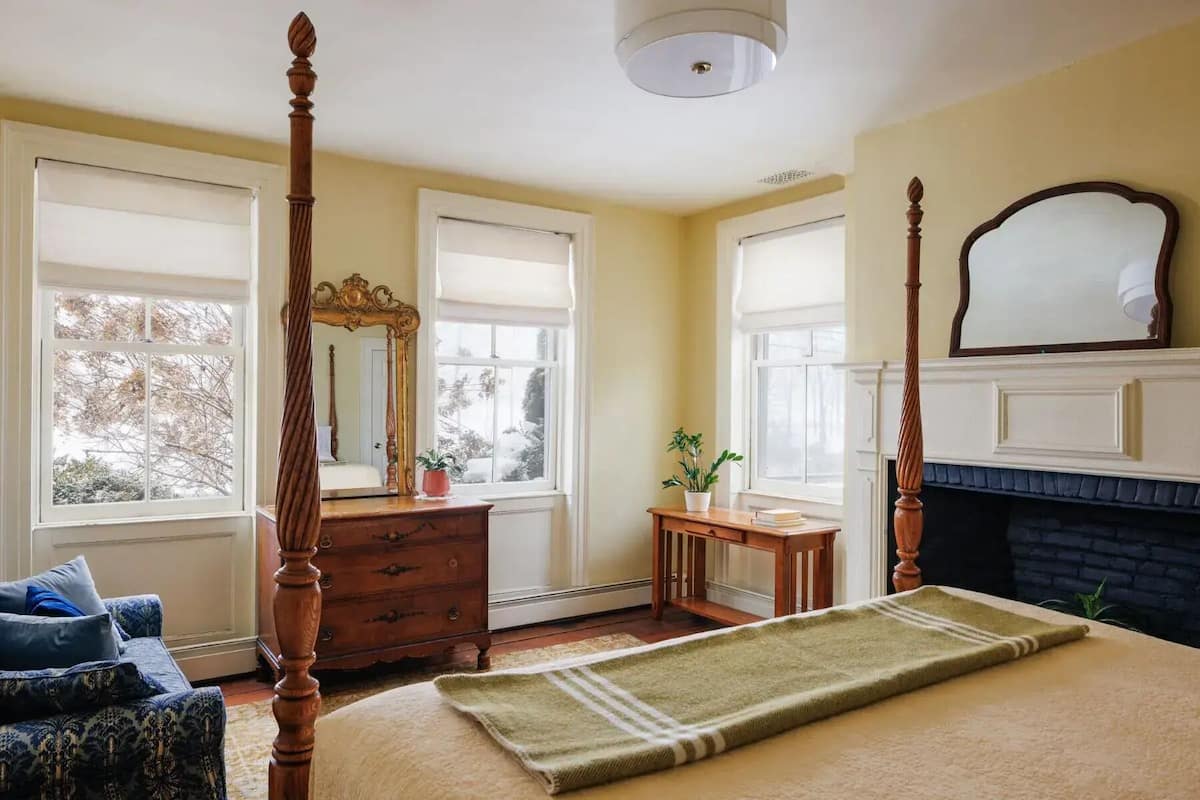 A bedroom with a four-poster bed, a dresser, a vanity with a mirror, a small table with a plant, and a fireplace.