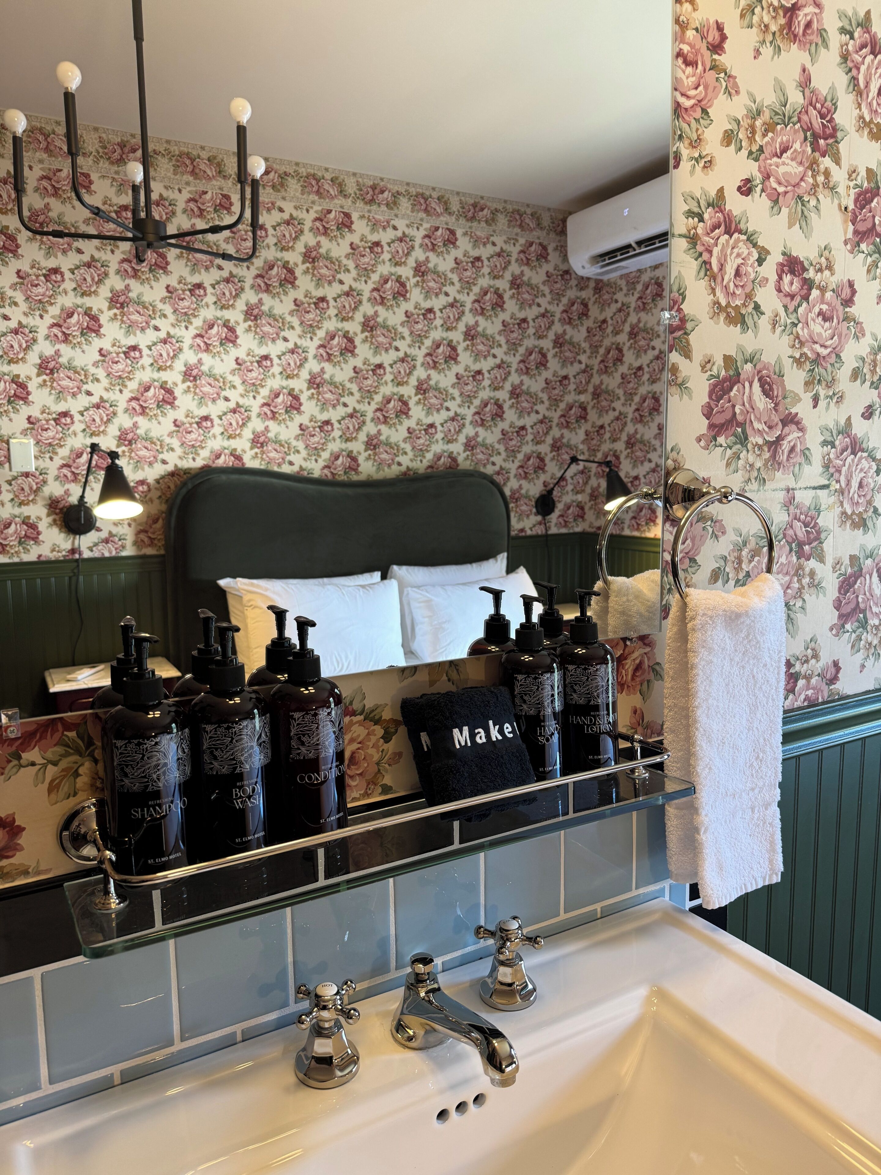 A bathroom with a floral wallpaper, a black shelf with toiletries, a white sink, and a towel rack.