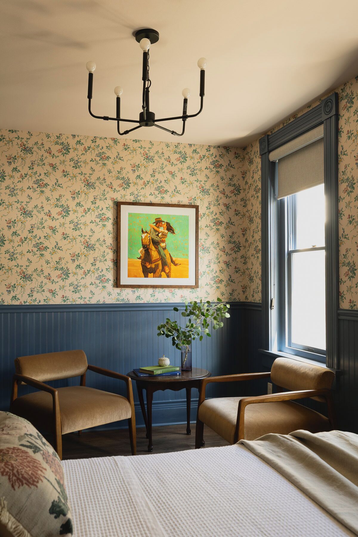 Una habitación con cama, dos sillas, una mesa con un florero, un cuadro y una ventana con vistas.