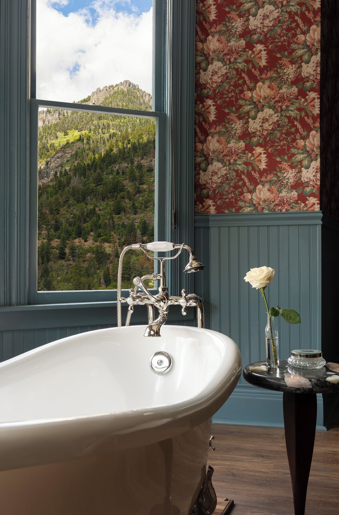 Un baño con una bañera independiente, una ventana con vistas a un paisaje montañoso y un papel tapiz floral.