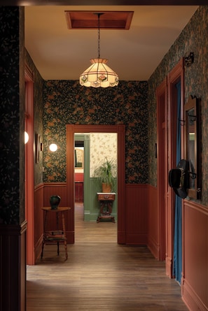 Un pasillo con papel tapiz estampado, suelo de madera, una lámpara colgante y una puerta que da a otra habitación.