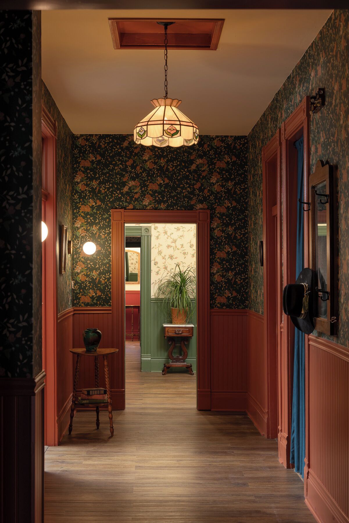 Um corredor com papel de parede estampado, piso de madeira, luminária pendente e uma porta que dá para outro cômodo.
