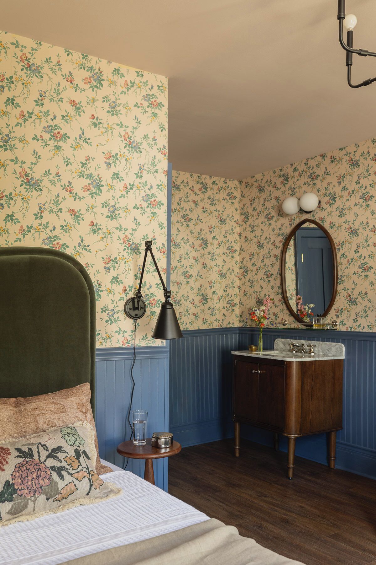 A room with a bed, a bedside table with a lamp, a dresser with a mirror, and a blue wall with floral wallpaper.