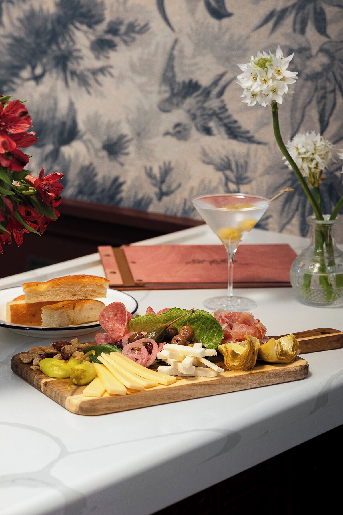 Una mesa con diversos alimentos, incluyendo pan, queso, aceitunas y carnes, acompañada de una copa de martini y un florero con flores.