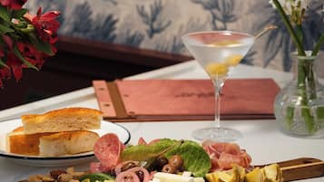 Una mesa con diversos alimentos, incluyendo pan, queso, aceitunas y carnes, acompañada de una copa de martini y un florero con flores.