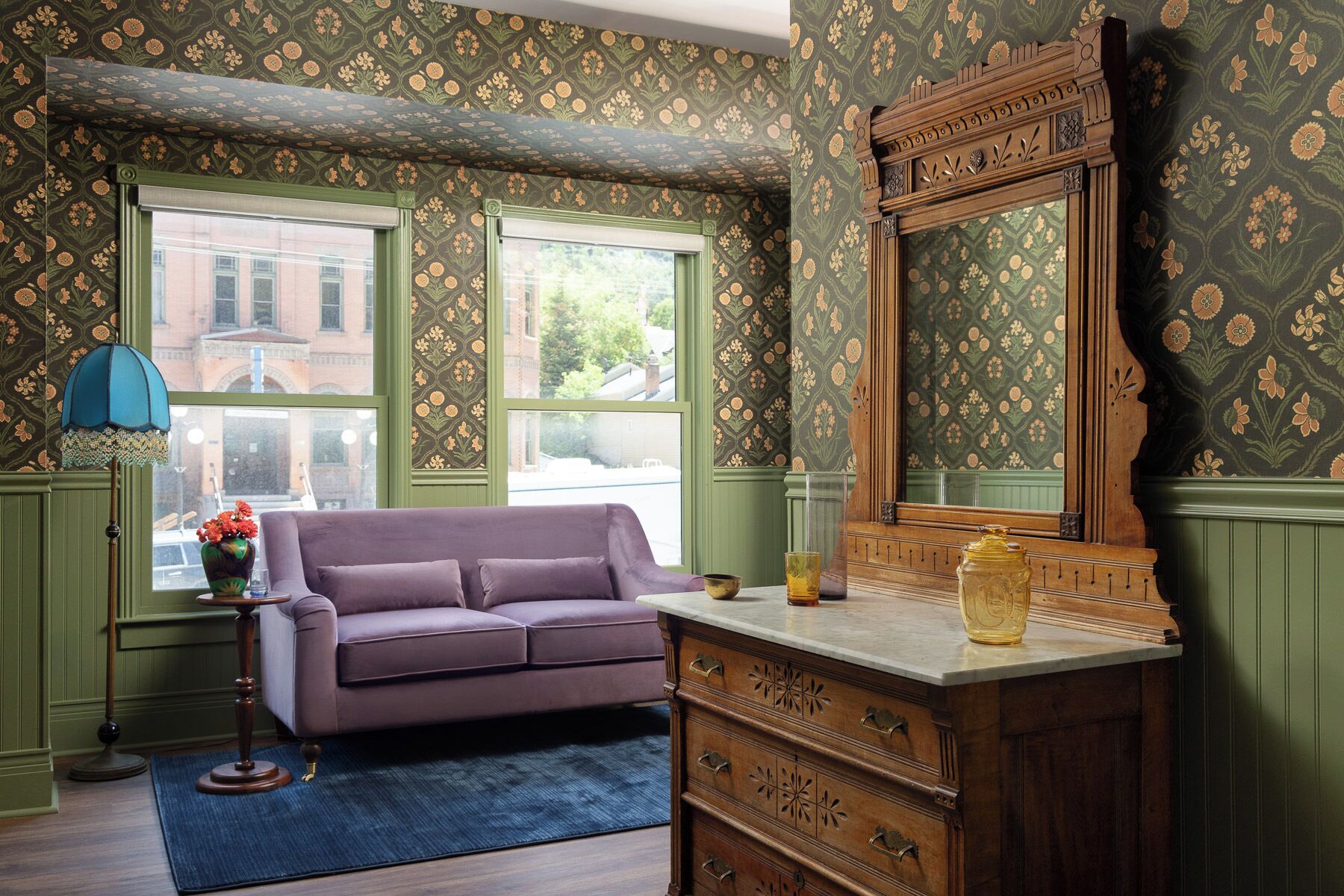A room with a purple sofa, a wooden dresser with drawers, a large mirror, and green patterned wallpaper.