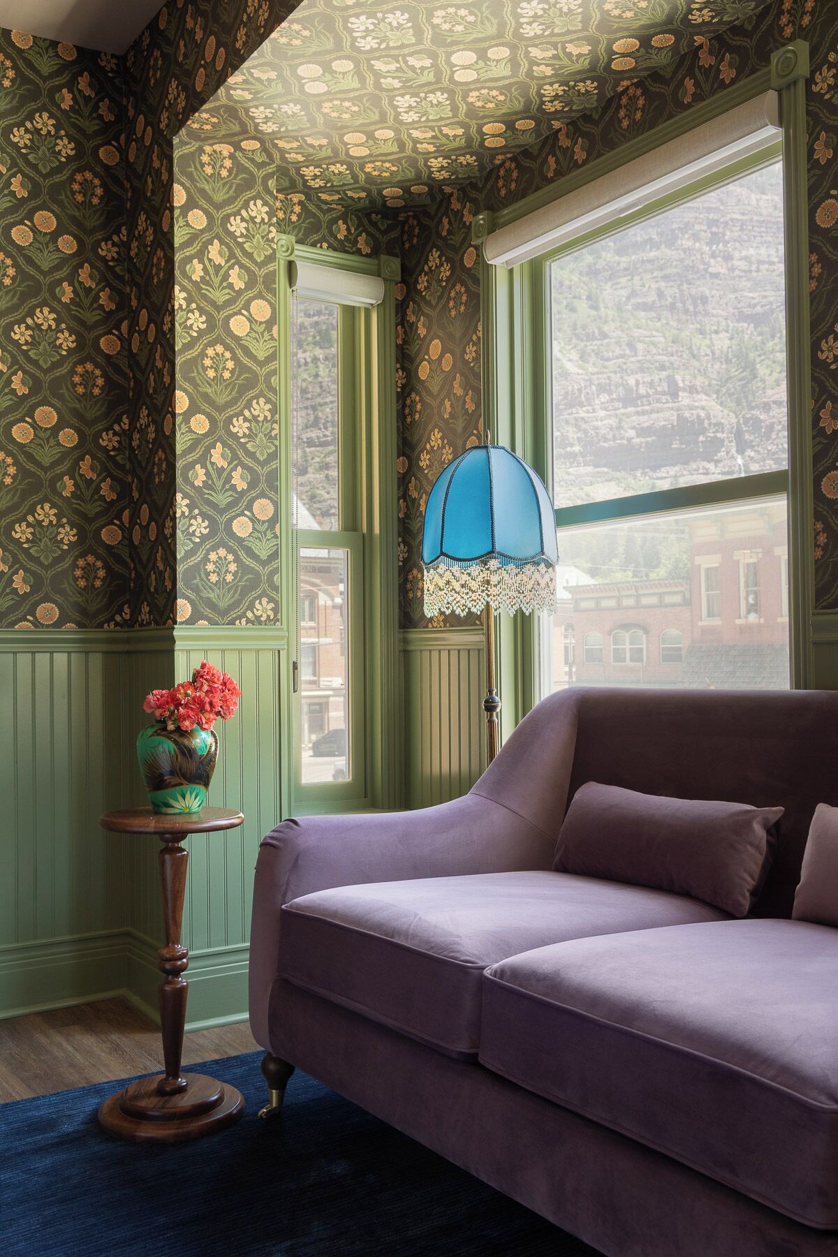 Una habitación con un sofá púrpura, un papel tapiz estampado en verde, un florero con flores y vistas a edificios a través de una ventana.