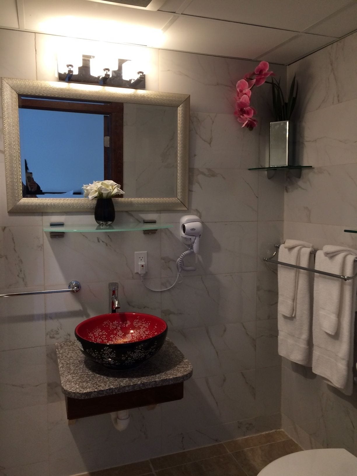 Un baño con pared de mármol, un lavabo redondo con un lavamanos rojo, un espejo sobre el lavabo, un perchero con toallas y una pequeña estantería con un jarrón de flores.