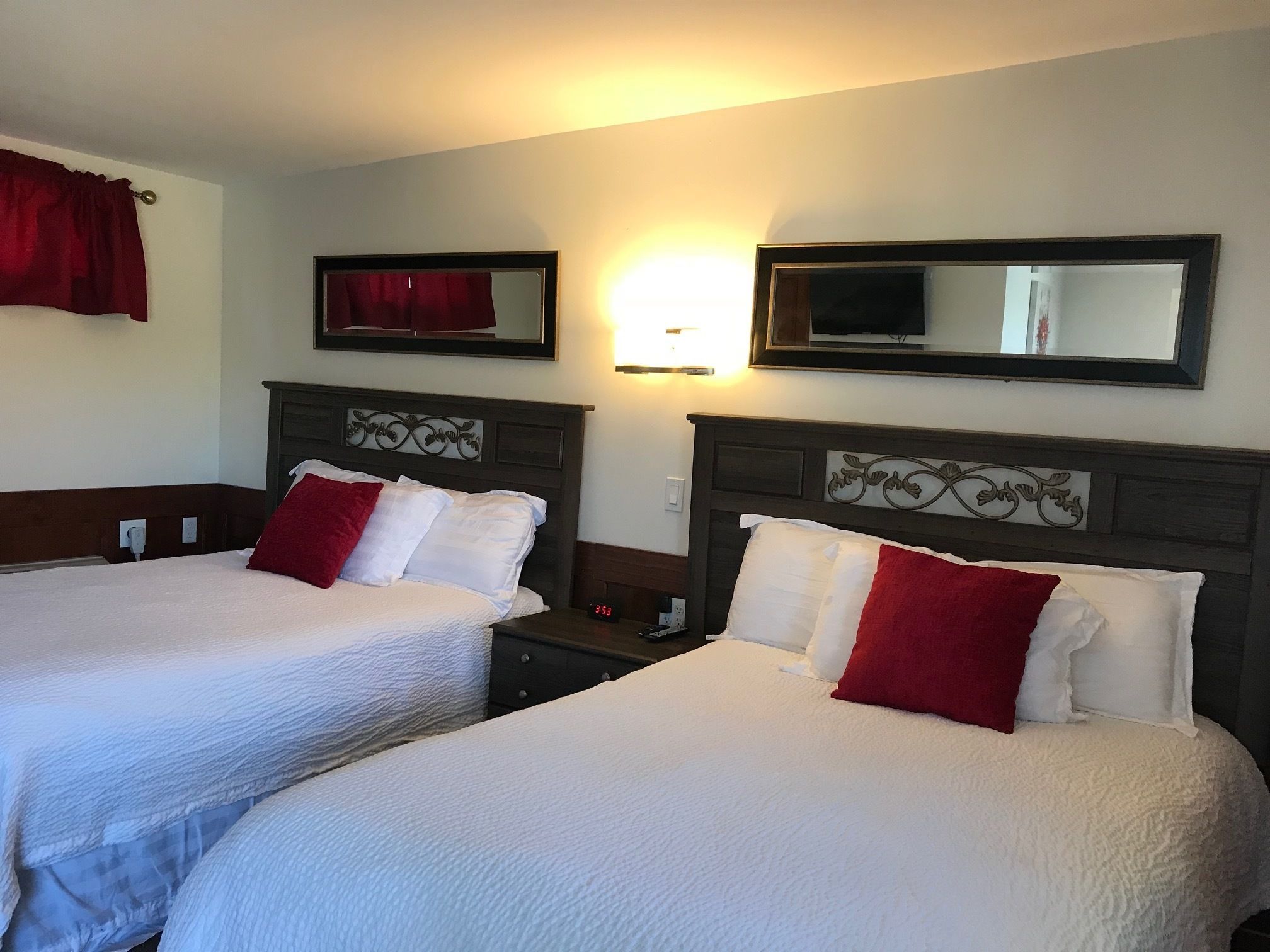 A hotel room with two beds, each with white bedding and red pillows, a nightstand with a clock, and a wall-mounted TV.